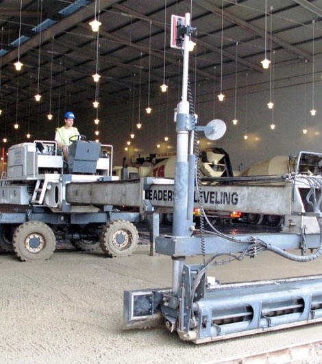 A worker operating a laser-guided concrete screeding machine in an indoor setting.