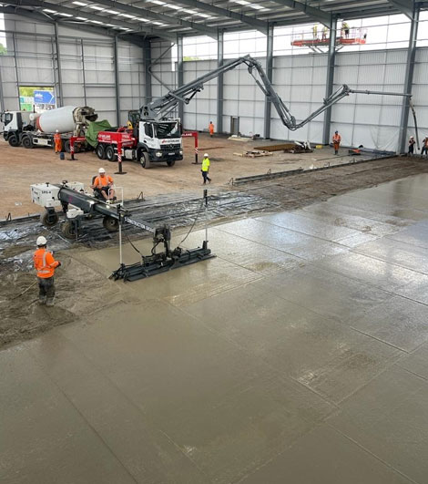 Workers using a concrete pump and screed to lay a smooth concrete floor in an industrial building.